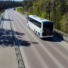 Biały autobus PKS Nova jedzie po moście wśród drzew.